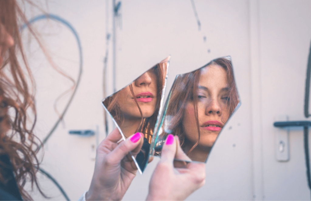 een vrouw die met 2 stukken van een gebroken spiegel in haar hand staat en waar je haar gezicht op 2 manieren in kan zien